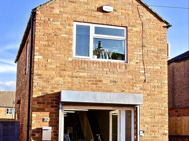 Claire Newberry's studio, with the two white doors open wide, shows the Shima Seiki & other knitting machines. The building is about 100 years old & is red brick built 3 storey high with an apex roof. The sky is blue behind the studio & the sun is shining brightly