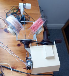 Image of a Knitmaster Yarn Twister. It is clamped to the table & by winding the handle two spools located on the rotating arms, spin round so the yarn twists around a core yarn making the candy cane effect on the yarn. Called a marl. There is yarn on the table which has come out of the twisting device. It's cream & brown in colour